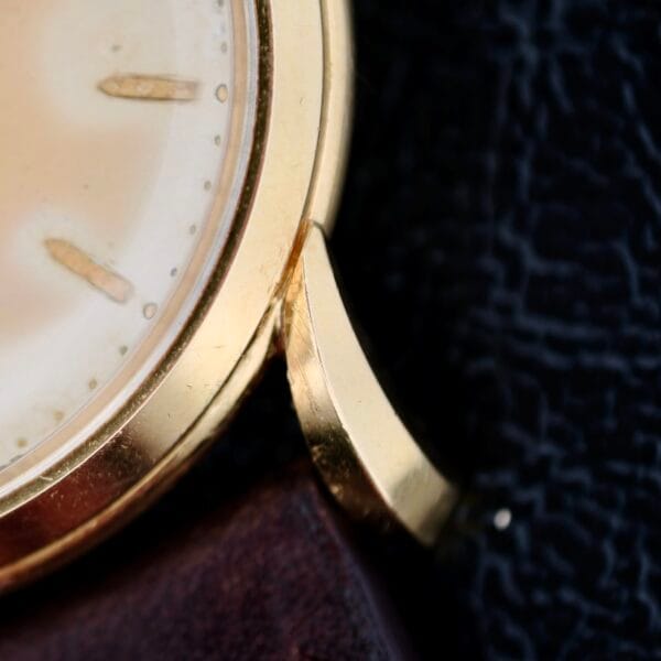 The Iwc Caltrava C. 89 features an 18k jumbo gold case connected to a brown leather strap. Its partially visible cream watch face with gold indices contrasts beautifully against a dark, textured background.