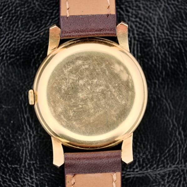 The IWC Caltrava C. 89 18k Jumbo Case, a vintage gold-colored watch back with noticeable scratches, features a brown leather strap with visible stitching on a textured black leather background.