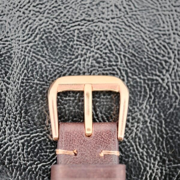 Close-up of the vintage Iwc Caltrava C. 89 18k Jumbo Case with a gold rectangular buckle against a textured black background, featuring curved edges and leather stitched with light thread.