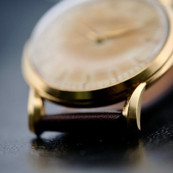 A vintage close-up of the Iwc Caltrava C. 89 18k Jumbo Case wristwatch with a brown leather strap on a reflective dark surface. The light-colored watch face, with gold hour markers, is partially blurred due to shallow depth of field, highlighting its classic metal casing and strap lugs.