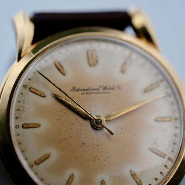 A close-up of the Iwc Caltrava C. 89 18k Jumbo Case wristwatch showcases its gold casing and aged, off-white dial. Delicate gold hour markers and hands add elegance, while the brand name is centrally printed, perfectly complemented by a dark leather strap.