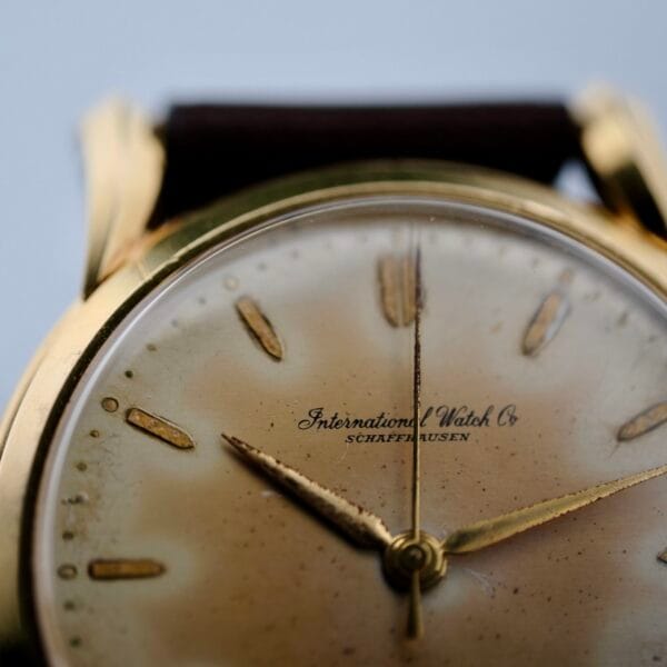Close-up of an Iwc Caltrava C. 89 18k Jumbo Case wristwatch with a faded gold-toned face, engraved International Watch Co Schaffhausen. It features simple hour markers and hands in a gold round case, its vintage charm highlighted by the partially visible leather strap.