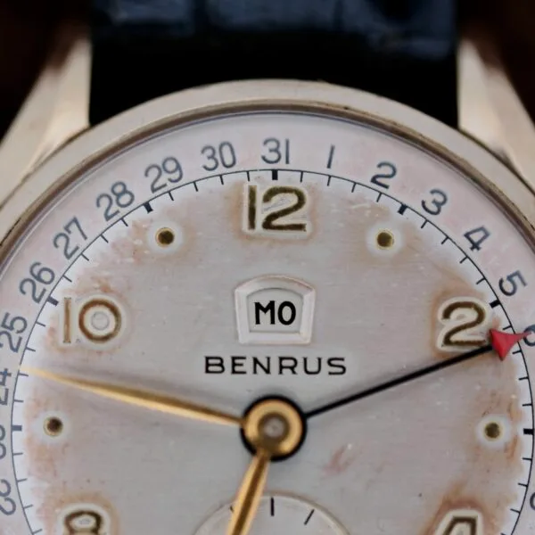 Close-up of a vintage Benrus Day/Date Pointer wristwatch featuring a circular dial with a date indicator. Gold hour, minute, and second hands adorn the face, while M0 sits above the brands name. The case displays a charming patina that enhances its timeless appeal.