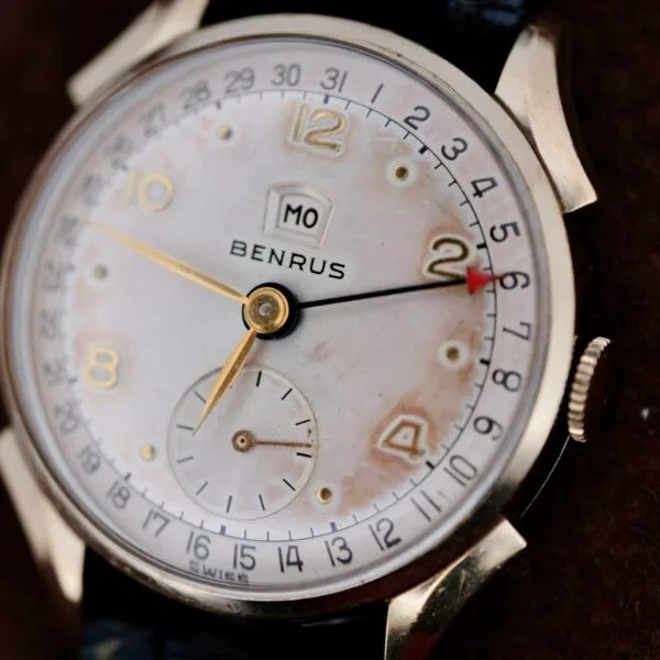Close-up of the vintage Benrus Day/Date Pointer wristwatch featuring a round face, gold hour markers and hands, a date window with MO, and a small subdial. Its gold casing paired with a leather band exudes timeless elegance.
