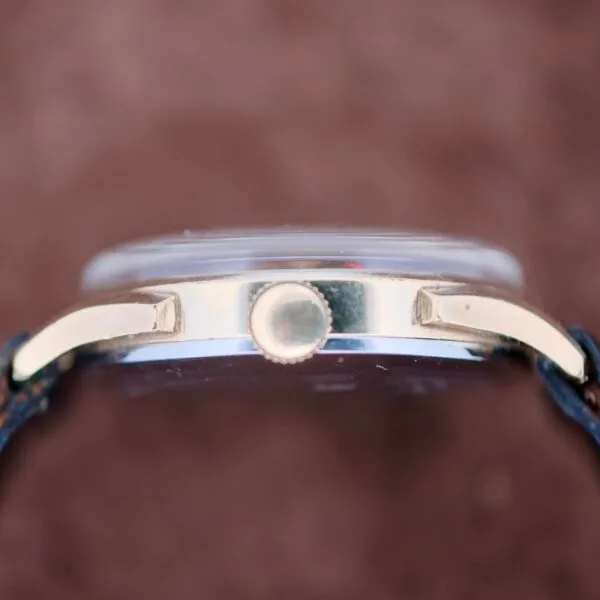 Side view of a vintage Benrus Day/Date Pointer wristwatch, showcasing a gold-colored case and center crown for time adjustment. It features a dark strap on a blurred brown surface.