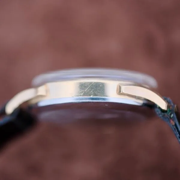 Side view of a Benrus Day/Date Pointer with a gold-tone case and black strap. The image focuses on the edge of the watch face and strap against a blurred brown background, showing a faint scratch on the metal surface.