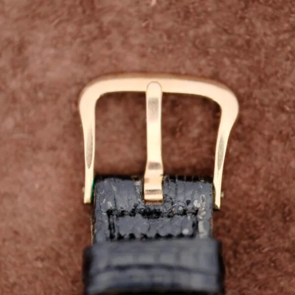 Close-up of a gold belt buckle, reminiscent of vintage craftsmanship, on a black leather strap with its intricate texture revealed against a brown textured background. The polished finish echoes the Benrus Day/Date Pointers classic allure.