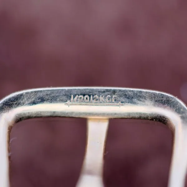 Close-up of a vintage Benrus Day/Date Pointer, featuring an engraved 1/20 12K G.F. marking. The metallic surface is worn and dusty against a blurred dark brown background.