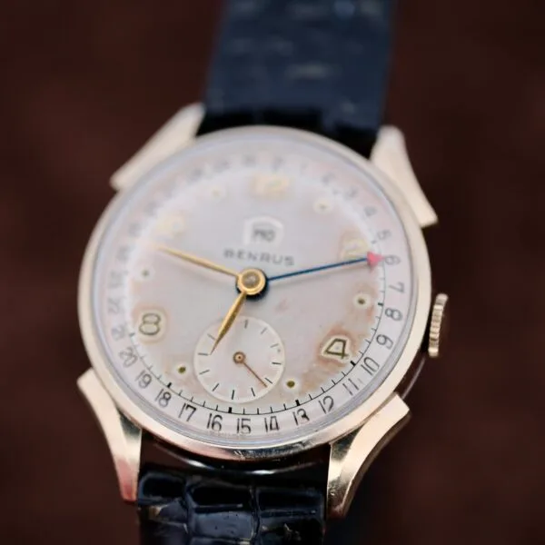 The Benrus Day/Date Pointer wristwatch features a gold case, black leather strap, and an elegant white dial with gold numbers. It includes a central date hand with a red tip and a small seconds subdial at 6 oclock, exuding timeless elegance.