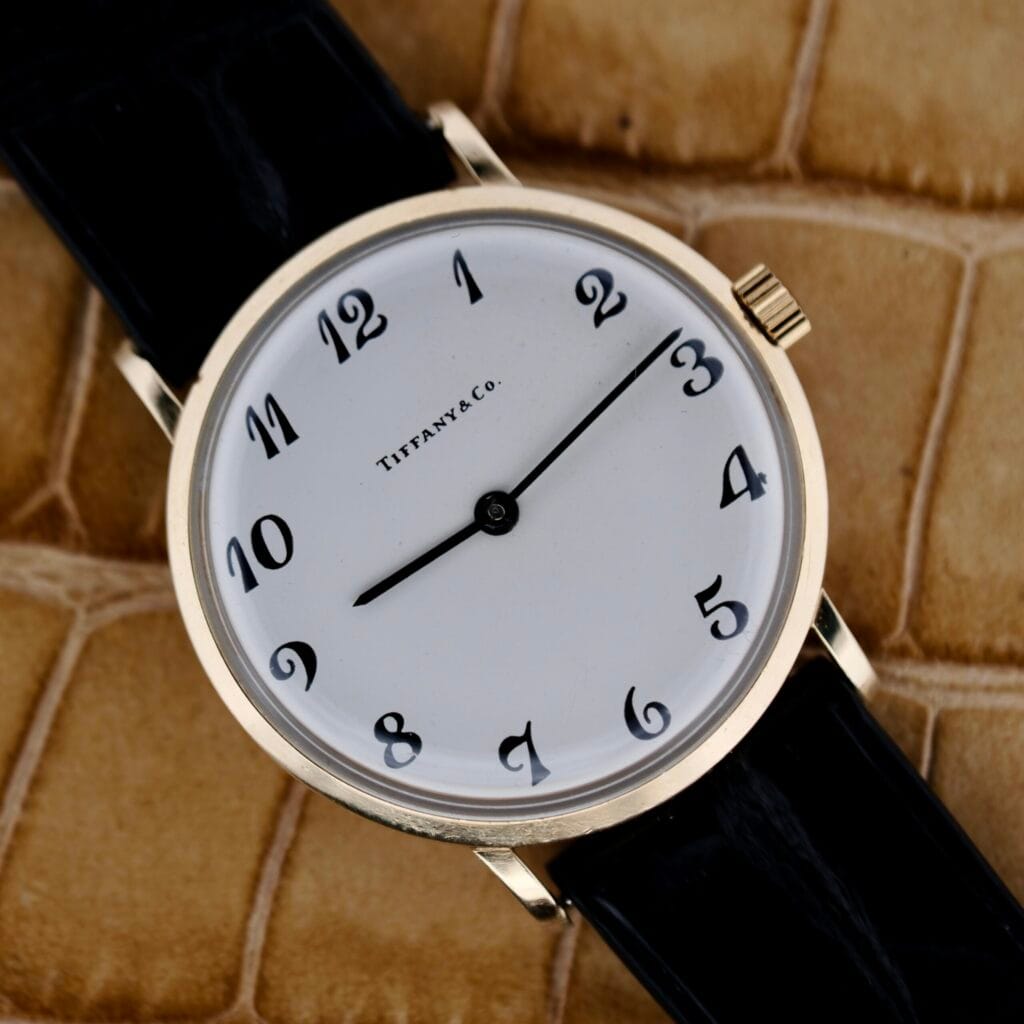 The Tiffany & Co Arabic Numerals Dial T-101 gold watch with a black strap rests on a tan textured background, elegantly showing 3:00.