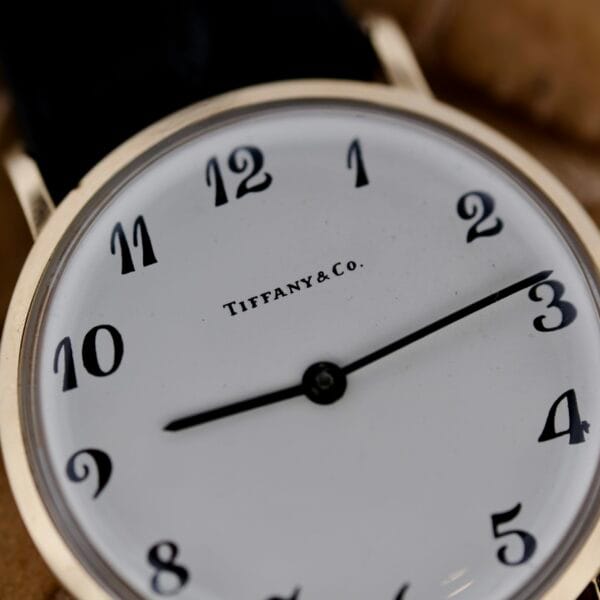 A close-up of the Tiffany & Co Arabic Numerals Dial T-101 wristwatch showcases its elegant details: a white face with classic black numerals and two hands marking time, all encased in a sleek metallic frame.