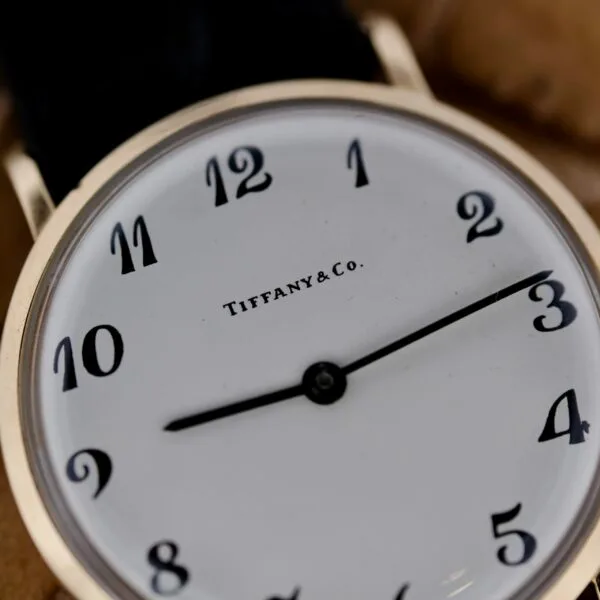 A close-up of the Tiffany & Co Arabic Numerals Dial T-101 wristwatch showcases its elegant details: a white face with classic black numerals and two hands marking time, all encased in a sleek metallic frame.