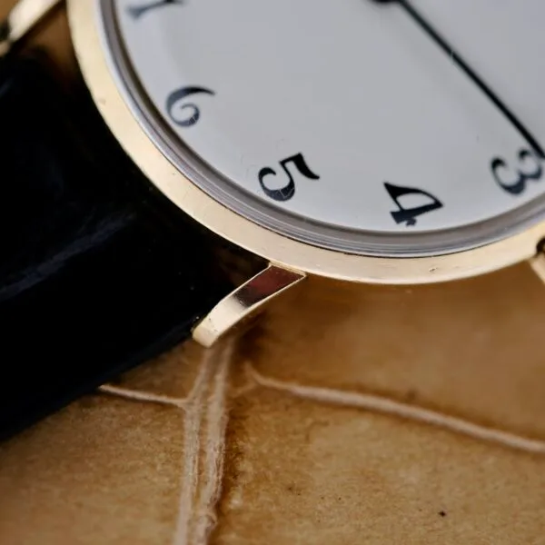 A close-up of the Tiffany & Co Arabic Numerals Dial T-101 showcases black numbers on a white face with a hint of a black leather strap. The unique numeral design adds charm to this elegant watch, resting gracefully on a light brown textured surface.