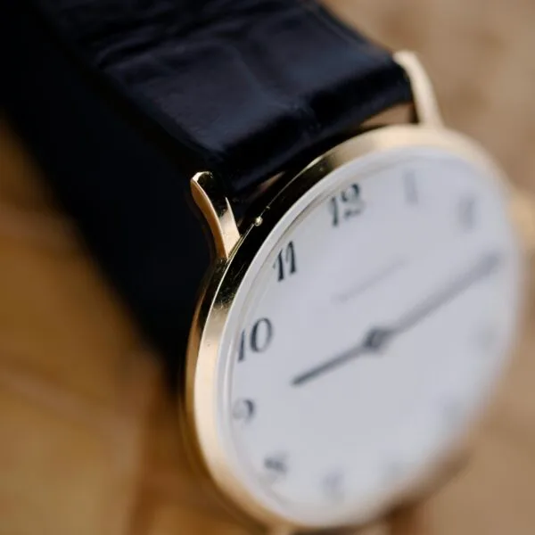 Close-up of a Tiffany & Co Arabic Numerals Dial T-101 wristwatch with a gold casing and black leather strap, exuding elegance. The watch features a white face with black numerals and thin black hands against a soft, blurry neutral background.