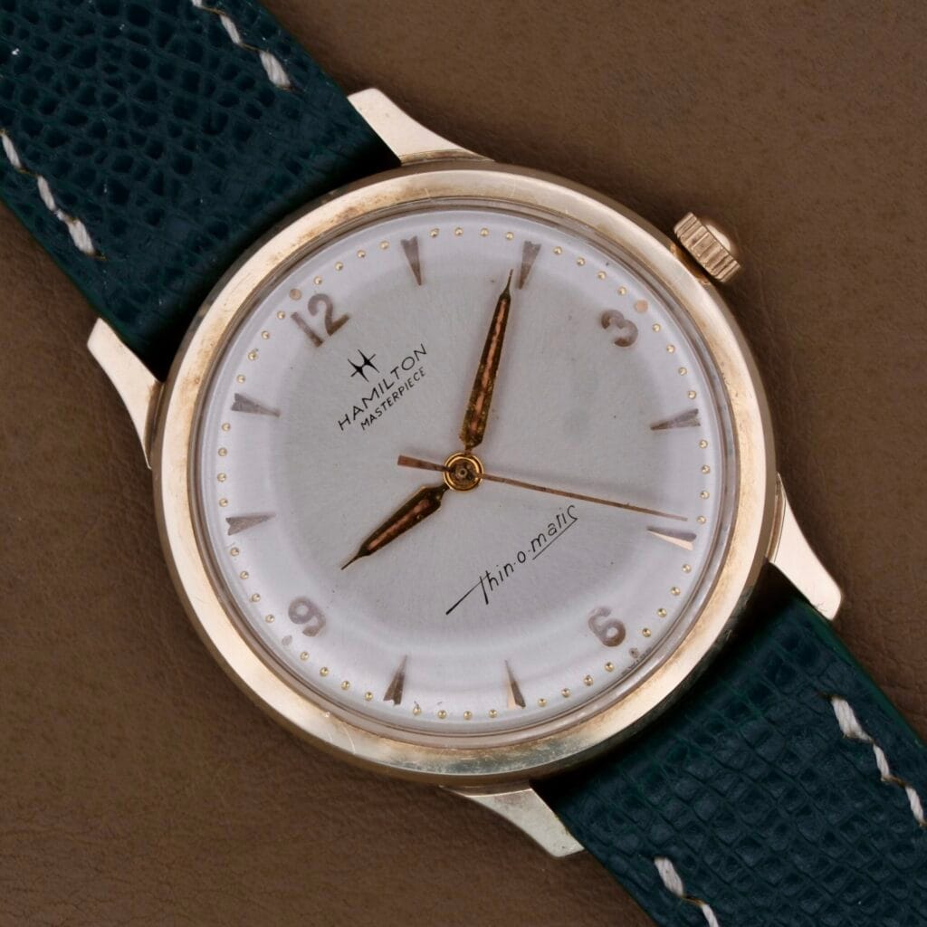 A Hamilton Masterpiece Thin-O-Matic 10k wristwatch with a white dial and green leather strap rests on a brown surface.