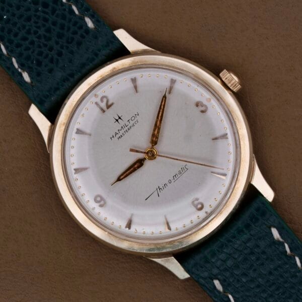 The Hamilton Masterpiece Thin-O-Matic 10k wristwatch boasts a gold-toned case and dial, dark green textured leather strap, gold hour markers, hands, and features Thin-o-Matic on the dial with a small inset second hand at six oclock.