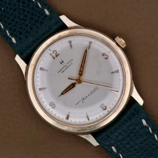 The Hamilton Masterpiece Thin-O-Matic 10k wristwatch boasts a gold-toned case and dial, dark green textured leather strap, gold hour markers, hands, and features Thin-o-Matic on the dial with a small inset second hand at six oclock.