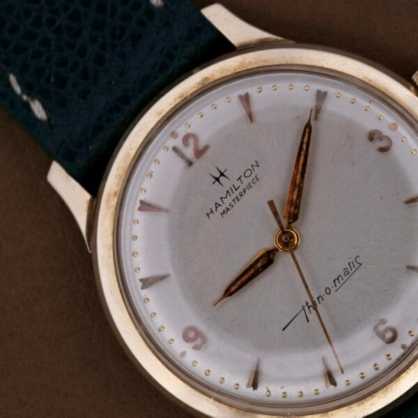 Close-up of the Hamilton Masterpiece Thin-O-Matic 10k wristwatch with a 10k gold accent and round face. Sleek gold hands move smoothly across the dial. Paired with a dark leather strap, it rests elegantly on a brown surface.