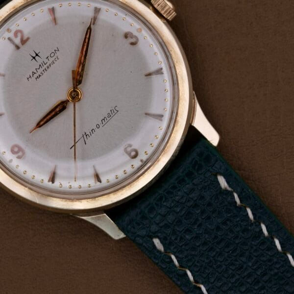A close-up of the Hamilton Masterpiece Thin-O-Matic 10k wristwatch showcases a white dial, gold hour markers, and Thin-o-matic text. It includes a blue leather strap with white stitching against a brown background.