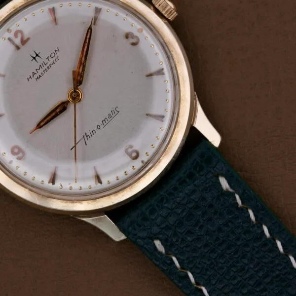 A close-up of the Hamilton Masterpiece Thin-O-Matic 10k wristwatch showcases a white dial, gold hour markers, and Thin-o-matic text. It includes a blue leather strap with white stitching against a brown background.