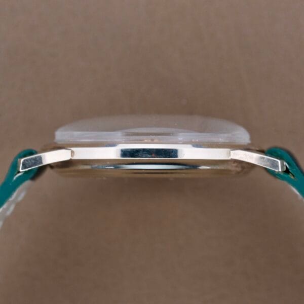 Side view of a Hamilton Masterpiece Thin-O-Matic 10k watch with a metal case and blue leather strap against a plain taupe background, embodying timeless elegance.