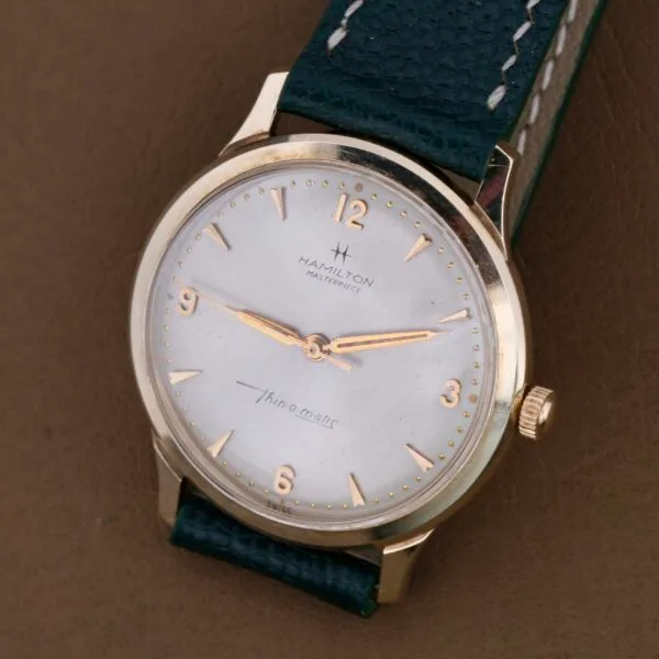The Hamilton Masterpiece Thin-O-Matic 10k is a vintage wristwatch with a gold-toned 10k case, black leather strap, and silver face. It features gold hour markers, hands, the brand name, and Thin-O-Matic text on a rich brown background, exuding timeless sophistication.