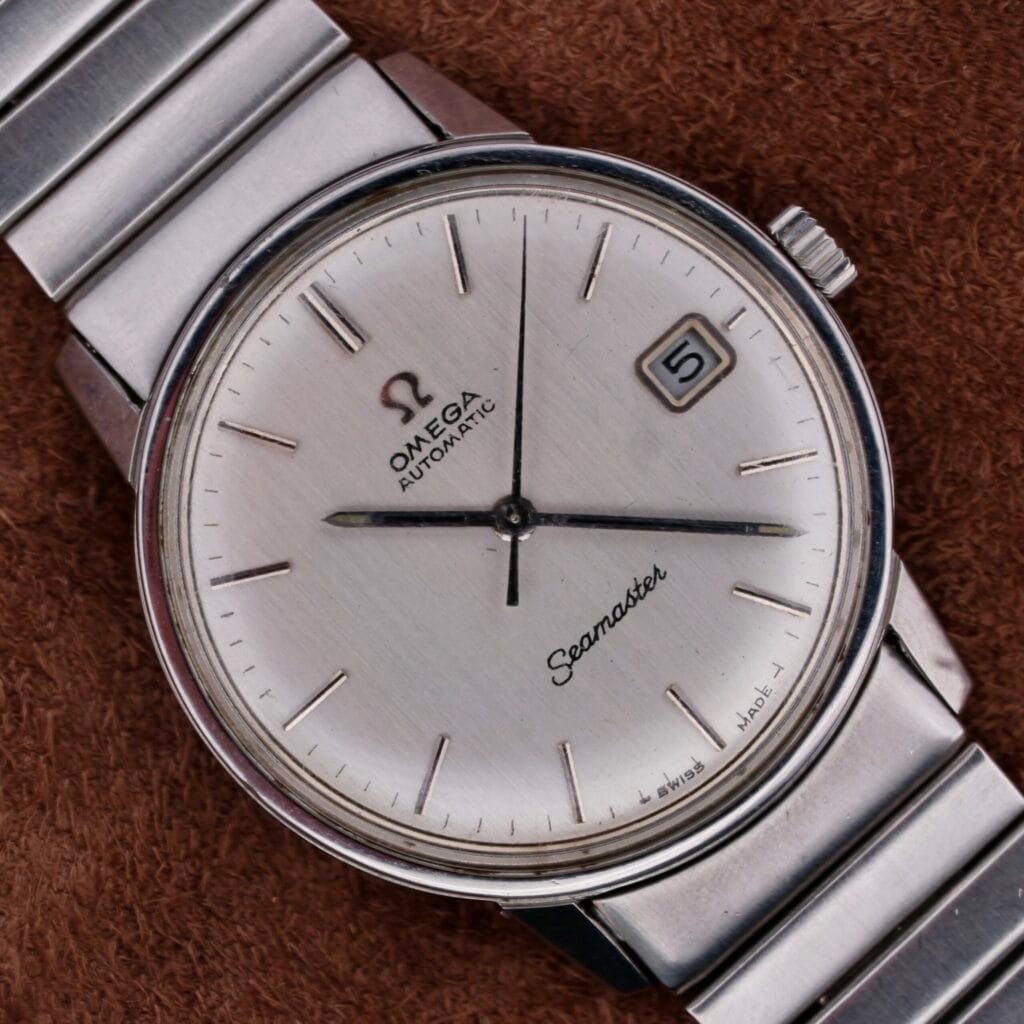 A close-up of the Omega Seamaster Automatic Date watch, featuring a silver dial and metal bracelet. It highlights automatic movement, simple hour markers, hands, and a date window at 3 oclock against a textured brown background.