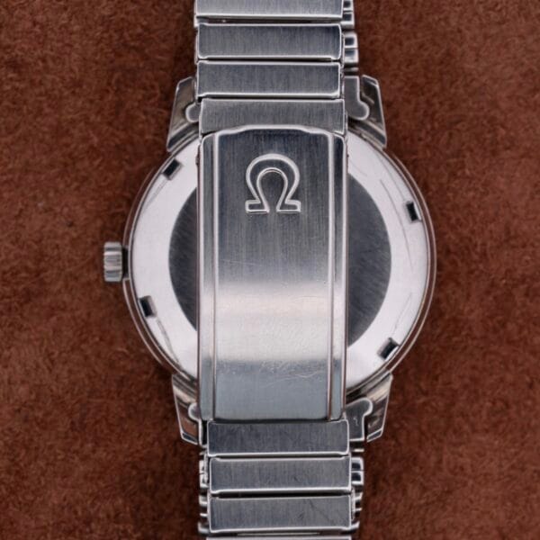 A close-up of the Omega Seamaster Automatic Dates silver band and case back, highlighting the iconic Omega logo on the clasp against a textured brown background, accentuating its metallic finish and automatic precision.