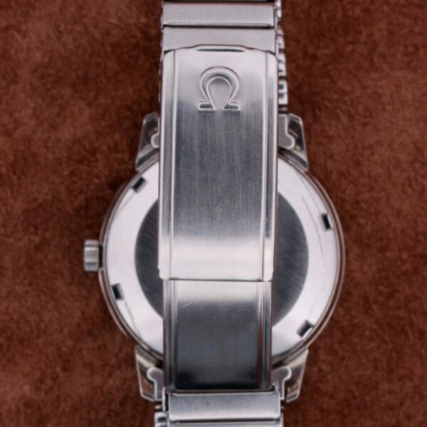Close-up of a stainless steel clasp engraved with the Omega Seamaster Automatic Date logo, resting elegantly on a soft, brown fabric background.