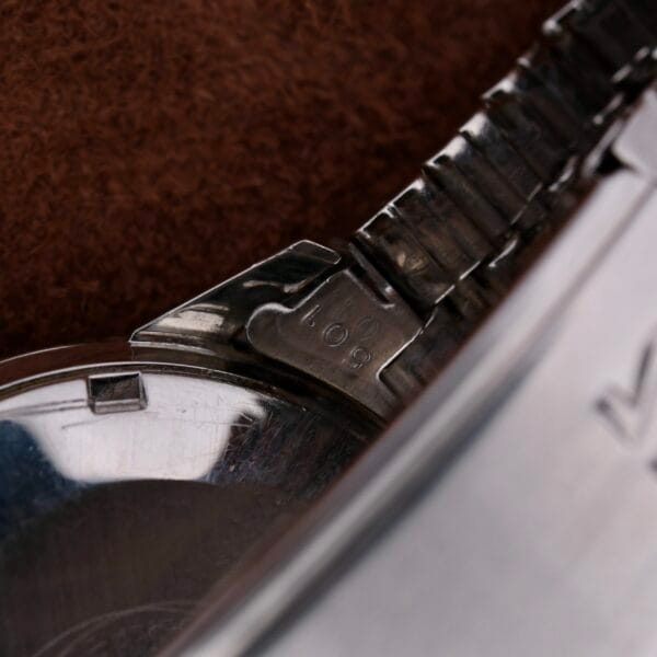 Close-up of a metallic wristwatch clasp with 108 engraved, from the Omega Seamaster Automatic Date. The watch face and part of the brown background are slightly visible, highlighting the intricate clasp and engraving details.