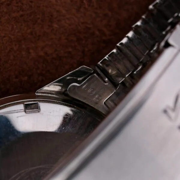 Close-up of a metallic wristwatch clasp with 108 engraved, from the Omega Seamaster Automatic Date. The watch face and part of the brown background are slightly visible, highlighting the intricate clasp and engraving details.