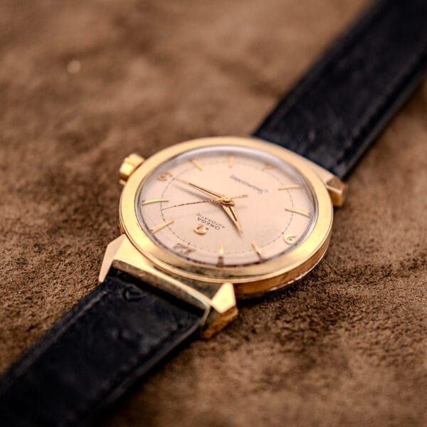 An Omega Seamaster Automatic Ref. 14350-2 Bull Horn Lugs gold wristwatch with a black leather strap is elegantly displayed on a brown suede surface.