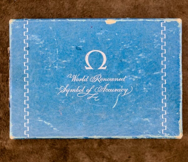 A worn blue Omega box with white text—World Renowned Symbol of Accuracy and the Omega logo—perfect for your prized Omega Seamaster Automatic Ref. 14350-2 Bull Horn Lugs.