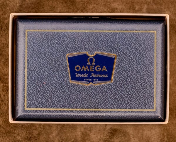 A vintage Omega watch box with a blue and gold logo and "World Famous Since 1848" on the lid—perfect for displaying your Omega Seamaster Automatic Ref. 14350-2 Bull Horn Lugs.