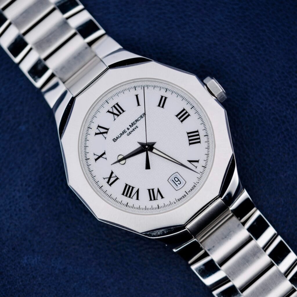 The Baume Mercier Riviera 65580 wristwatch features a white face with Roman numerals and date display, all set against a dark blue textured background.