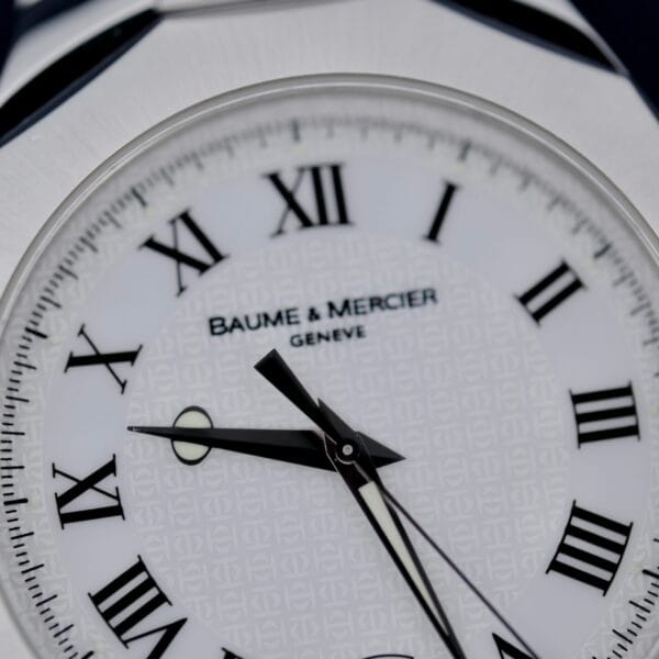 Close-up of a Baume Mercier Riviera 65580 watch face featuring Roman numerals, a patterned white background, black hour and minute hands, and the brand name inscribed below XII numeral.