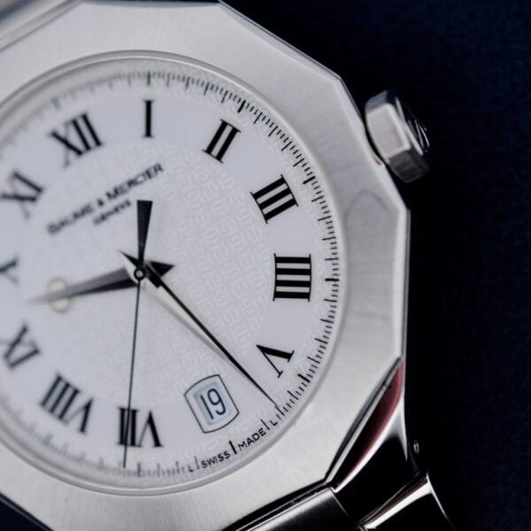 A close-up of the Baume Mercier Riviera 65580 wristwatch showcases its white face with Roman numerals, date window set to the 19th, silver hour and minute hands, and a polished hexagonal bezel. The dial features the brand name and Swiss Made text.