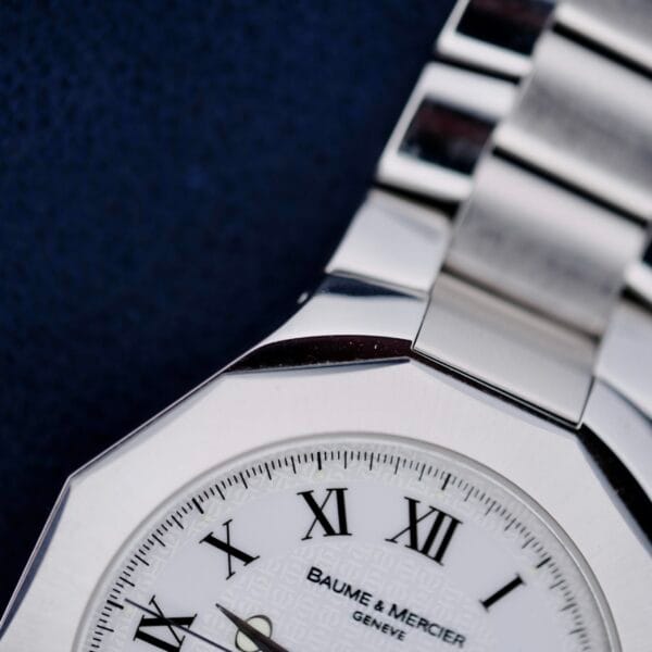 Close-up of a Baume Mercier Riviera 65580 watch face featuring Roman numerals and a white dial. A portion of the silver metal band is visible, with Baume & Mercier Genève printed on the dial against a dark, textured background.