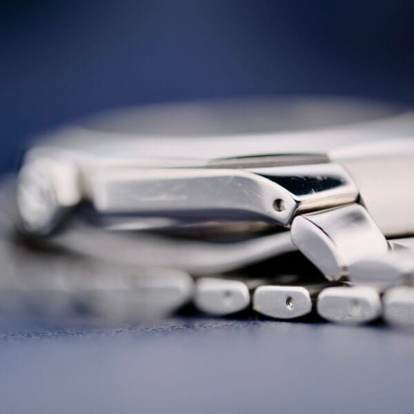 A close-up of the Baume Mercier Riviera 65580 reveals a side view of its metallic bracelet and polished silver case edges, with a blurred blue background.