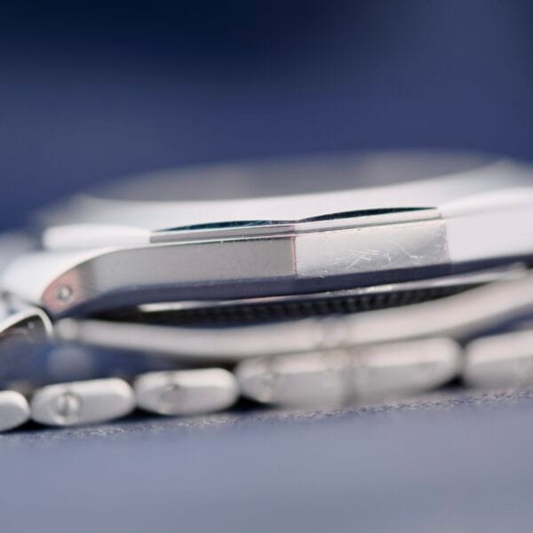 Close-up of a Baume Mercier Riviera 65580 stainless steel wristwatch viewed from the side, highlighting the watch case and band links against a blurred dark background, with minor scratches and wear on the surface.