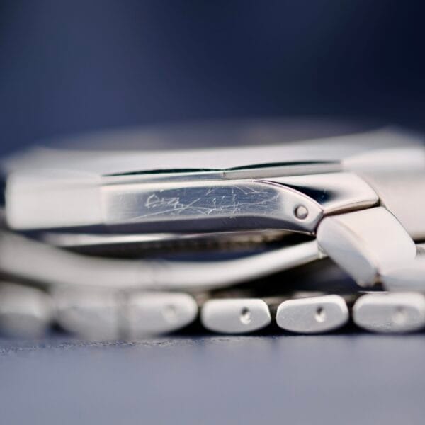 Close-up image of the silver bracelet of a Baume Mercier Riviera 65580, displaying fine scratches on the clasp. The watch rests on a smooth surface with a blurred soft blue-gray background.