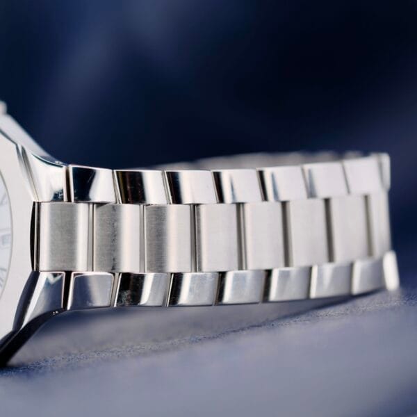 Close-up of the Baume Mercier Riviera 65580 stainless steel bracelet on a dark blue textured background. The bracelets alternating polished and brushed links highlight its blend of shiny and matte finishes.