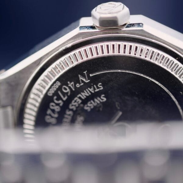 A close-up of the Baume Mercier Riviera 65580 wristwatch reveals engraved text with numbers and Swiss on the back. The edge features a grooved pattern, and part of the metal bracelet is in view. The watch crown is prominent on the side.
