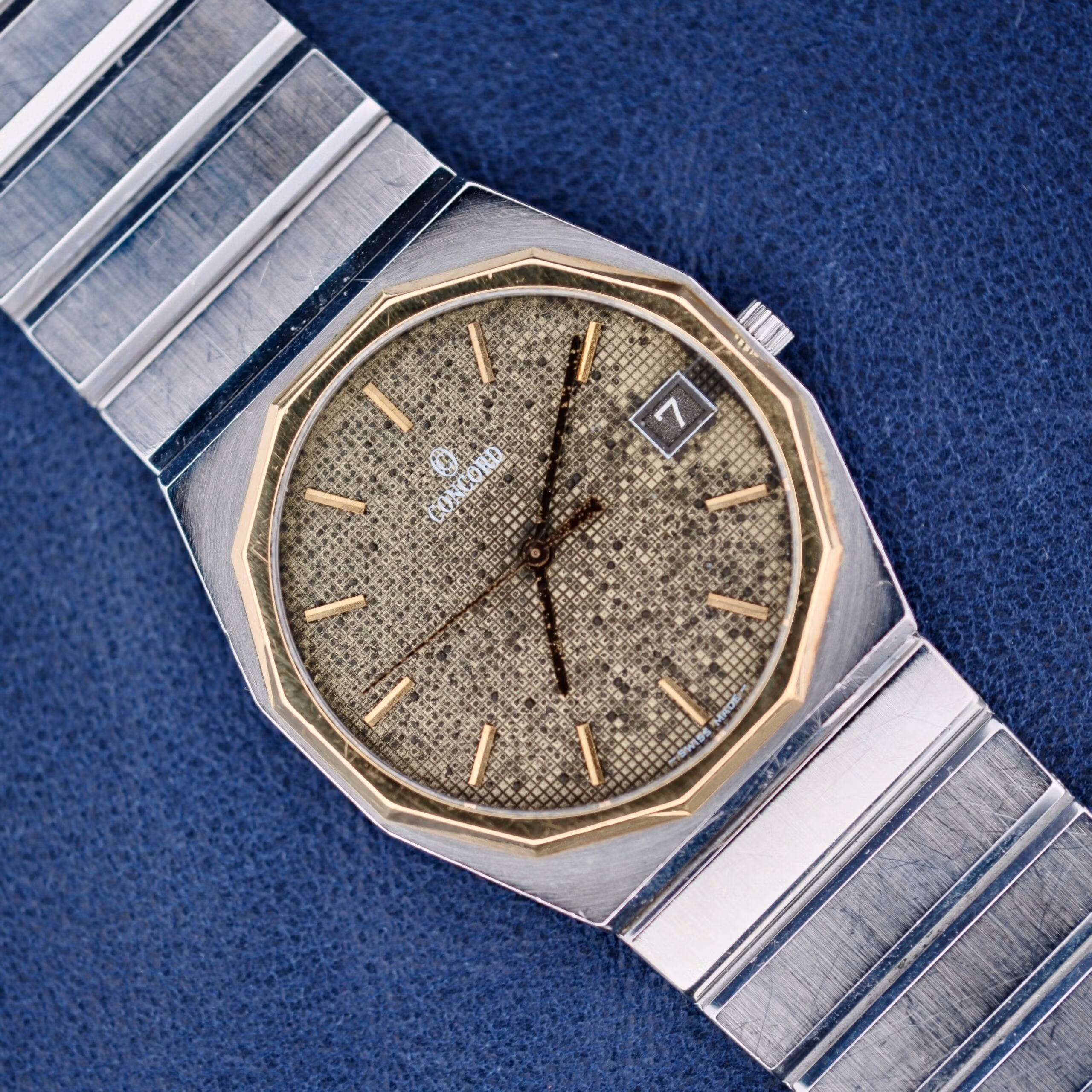 Concord Mariner SG with 18k gold bezel, textured silver dial, gold markers, and date window displayed against a blue background.