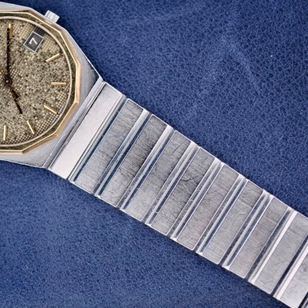 A close-up of the Concord Mariner SG wristwatch, featuring a textured silver round face, date display window, and metallic band made of rectangular links. The watch rests on a blue textured surface.