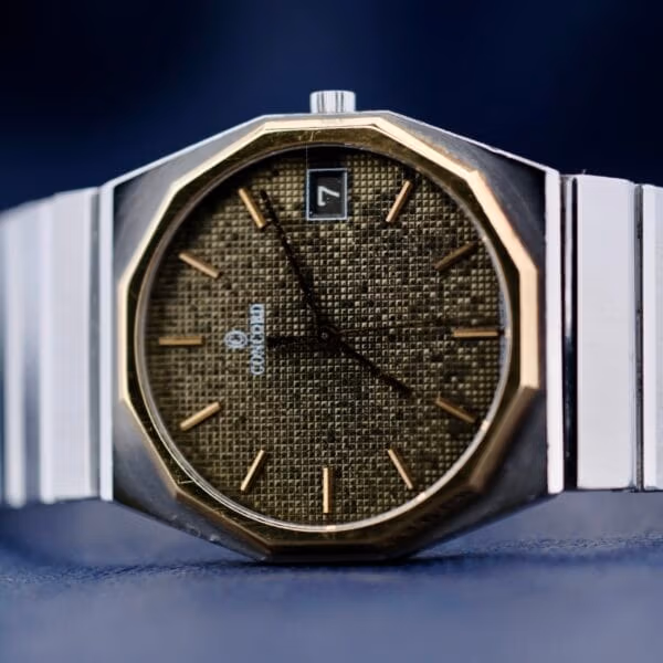 The Concord Mariner SG 18k Gold Bezel features a wristwatch with a metallic silver band and a textured dark brown face, gold hour markers and hands showing about 10:10, and a small date window at the 3 oclock position.