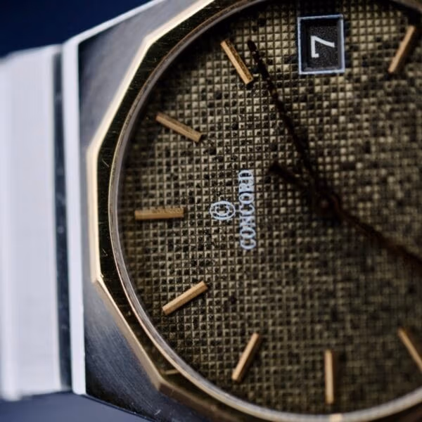 Close-up of the luxury Concord Mariner SG 18k Gold Bezel watch with a textured brown face, thin gold hour markers and hands, and a centered brand name. A date window shows the number 7, complemented by a metal bezel and band.