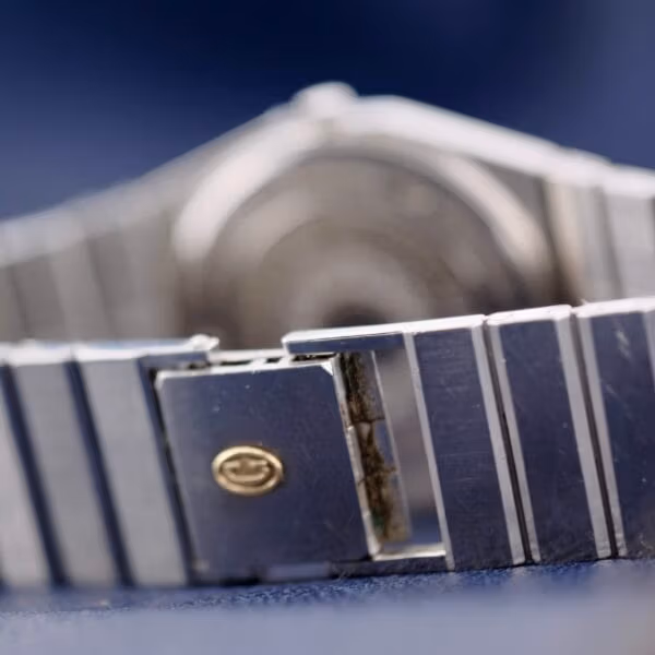 Close-up of a metallic watch band featuring horizontal grooves and a gold emblem on the clasp. The Concord Mariner SG 18k Gold Bezel is slightly out of focus, with a blue surface beneath the band.