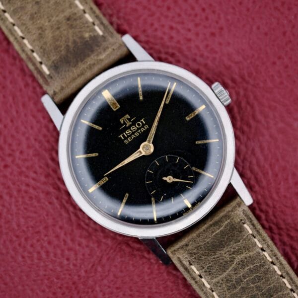 The Tissot Seastar Black Gilt Dial Sub Second is displayed with its black dial, gold hour markers, small second subdial, and leather strap on a textured red surface.