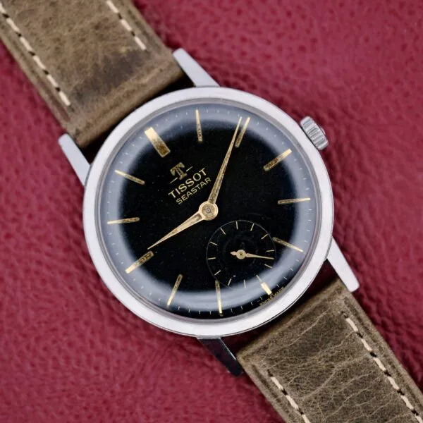 The Tissot Seastar Black Gilt Dial Sub Second is displayed with its black dial, gold hour markers, small second subdial, and leather strap on a textured red surface.